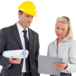 White businessman in a yellow hard hat holding a roll of architectural drawings while a blonde white woman shows him something on a clipboard