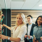 White woman with blonde hair placing sticky notes on glass wall while colleagues watch