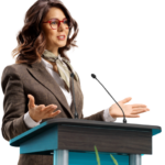 White woman with brown hair and red glasses in brown suit jacket giving a speech at a podium